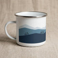 A white enamel mug with a silver rim featuring a blue mountain landscape graphic and two bicycles.