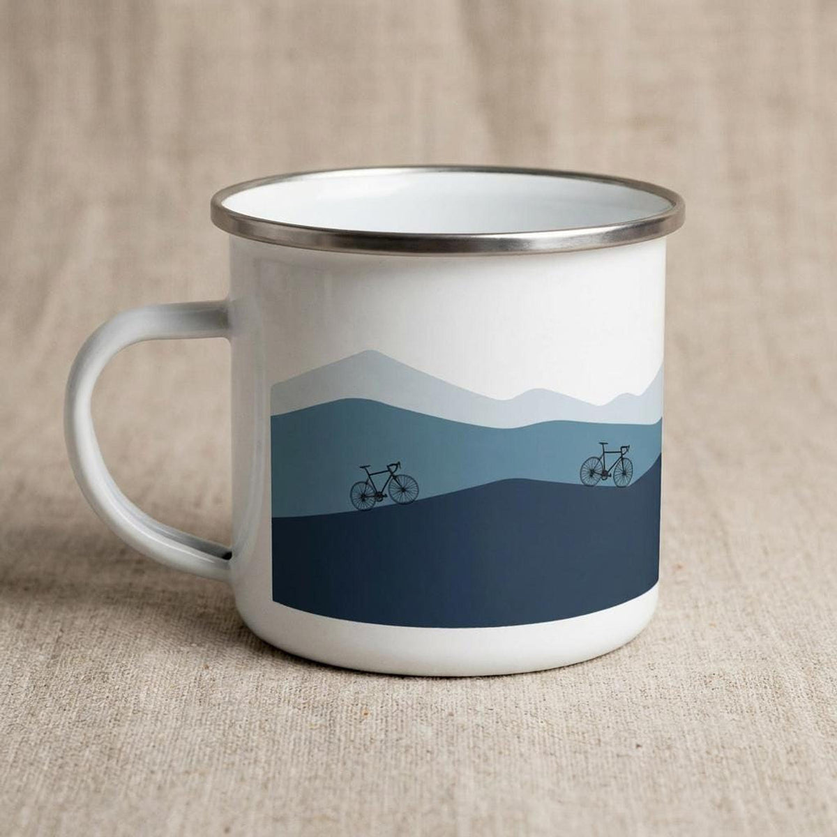A white enamel mug with a silver rim featuring a blue mountain landscape graphic and two bicycles.
