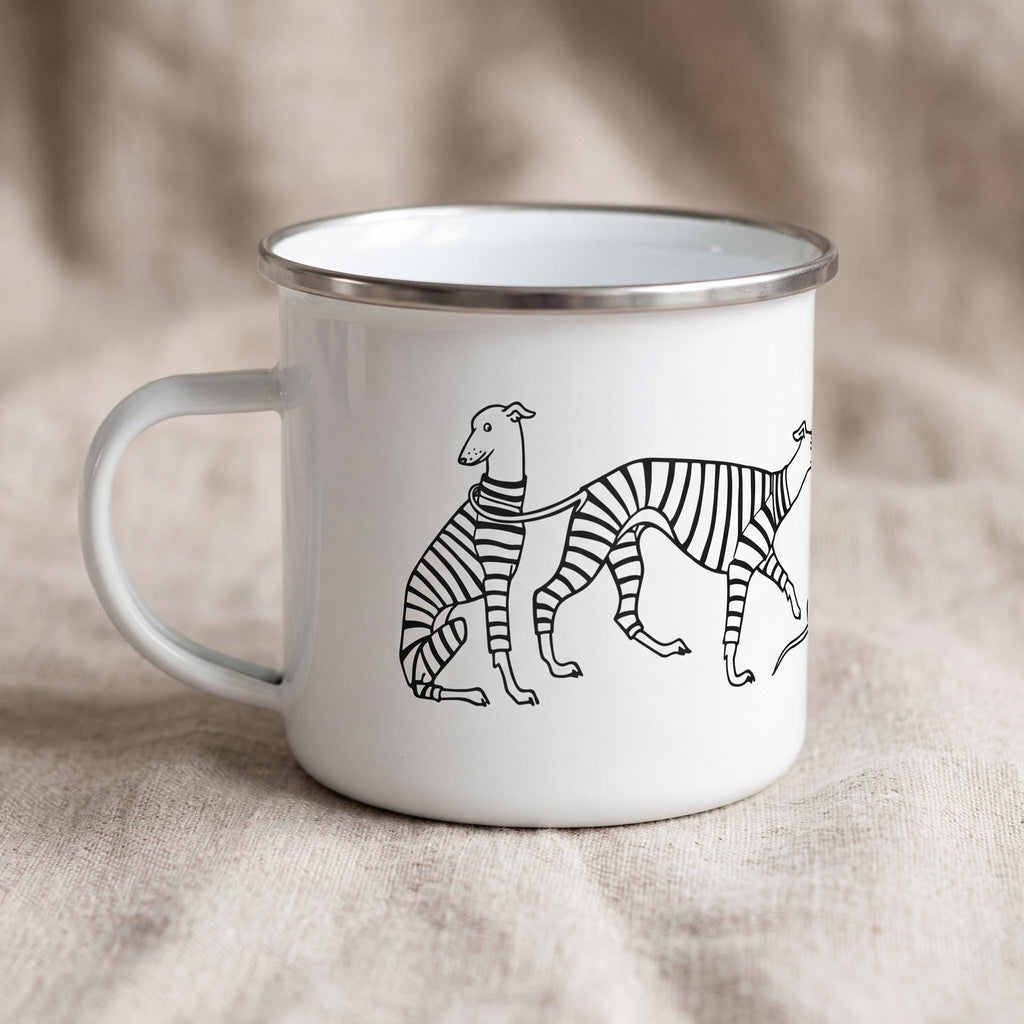 A white enamel mug with a silver-toned rim and a handle on the left side. The mug features a black line drawing of two greyhound-like dogs wearing striped sweaters, connected by a leash, printed on the front of the white surface.