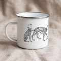 A white enamel mug with a silver-toned rim and a handle on the left side. The mug features a black line drawing of two greyhound-like dogs wearing striped sweaters, connected by a leash, printed on the front of the white surface.