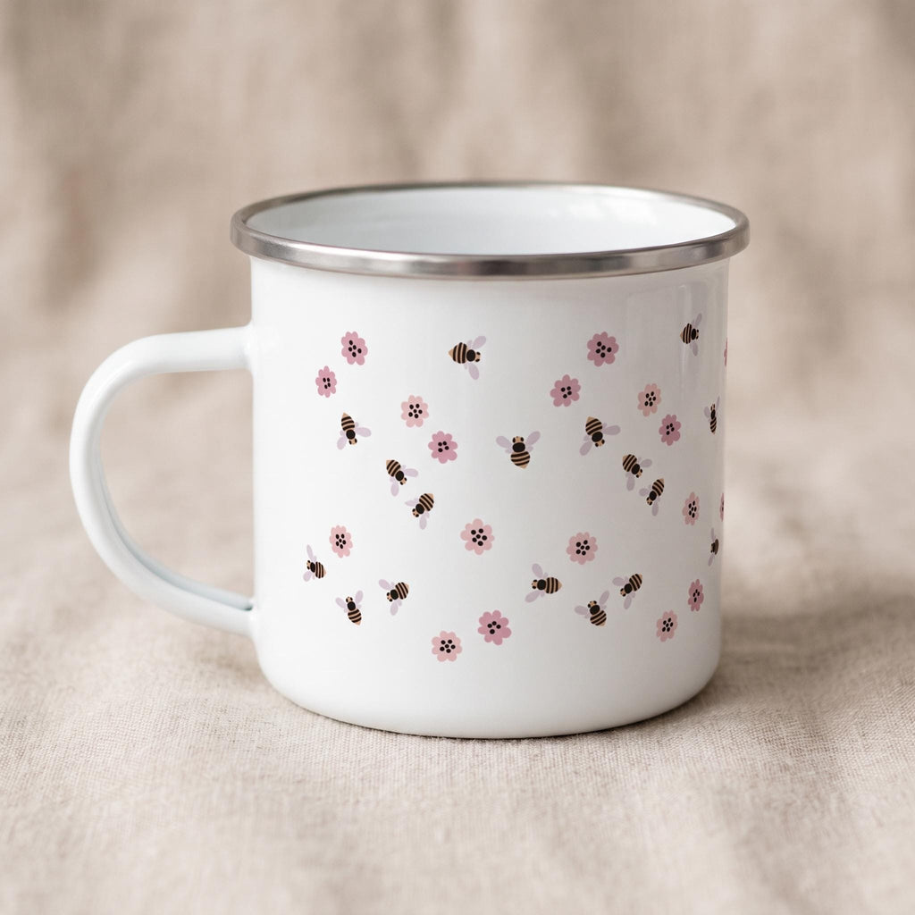 A white enamel mug featuring a repeating pattern of small brown and black bees interspersed with tiny pink flowers. The mug has a smooth white finish, a curved handle on the left side, and a polished silver-tone metal rim at the top edge.