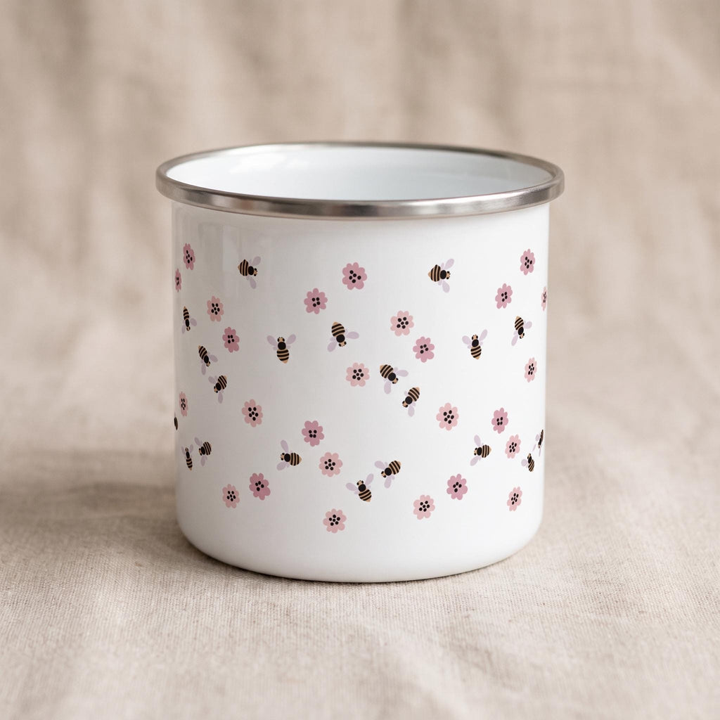 A white enamel mug featuring a repeating pattern of small pink flowers and brown and black striped bees. The mug has a smooth, cylindrical body and a silver-coloured metal rim around the top edge, providing a clean and simple aesthetic.