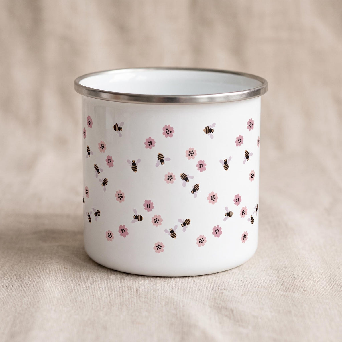 A white enamel mug featuring a repeating pattern of small pink flowers and brown and black striped bees. The mug has a smooth, cylindrical body and a silver-coloured metal rim around the top edge, providing a clean and simple aesthetic.