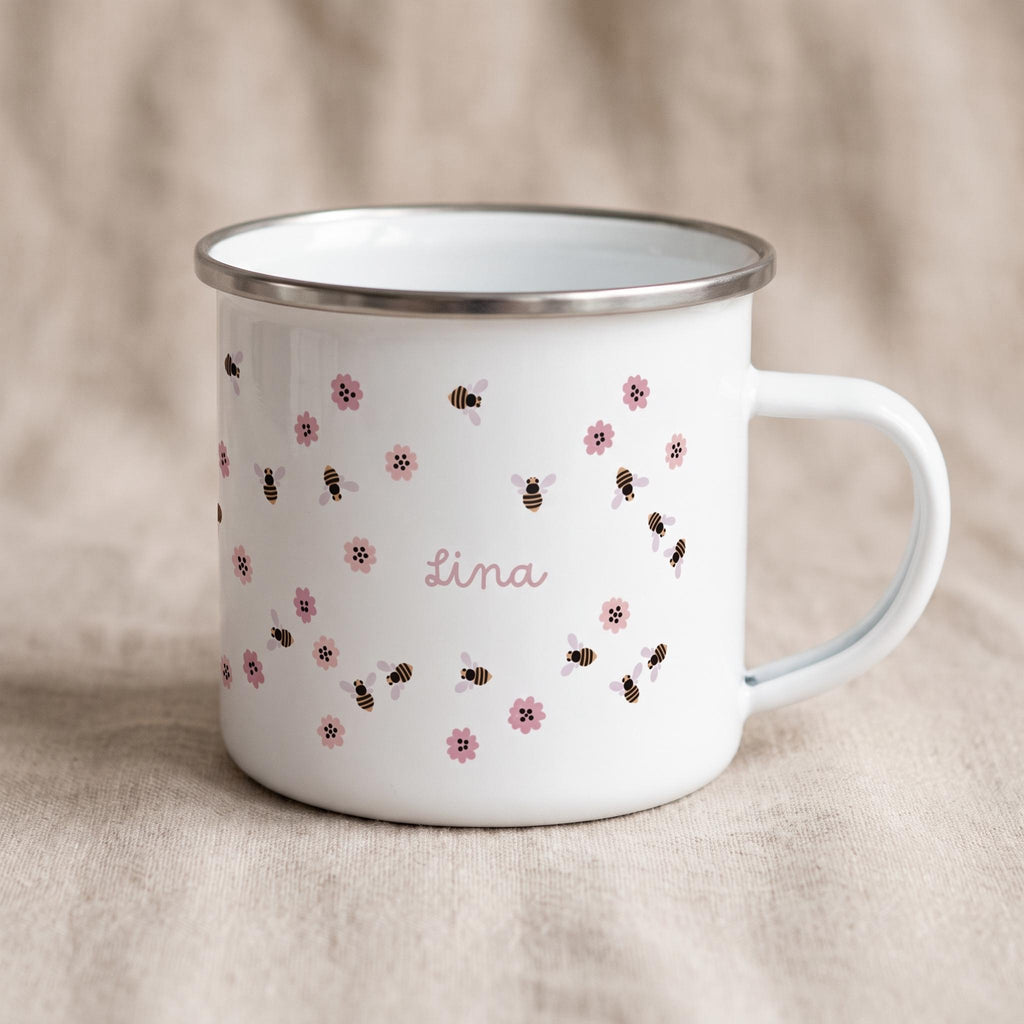 A white enamel mug with a silver rim and a handle, featuring a repeating pattern of small brown and black bees and pink flowers. The name Lina is printed in a pink script font in the centre of the mug among the bee and flower design.