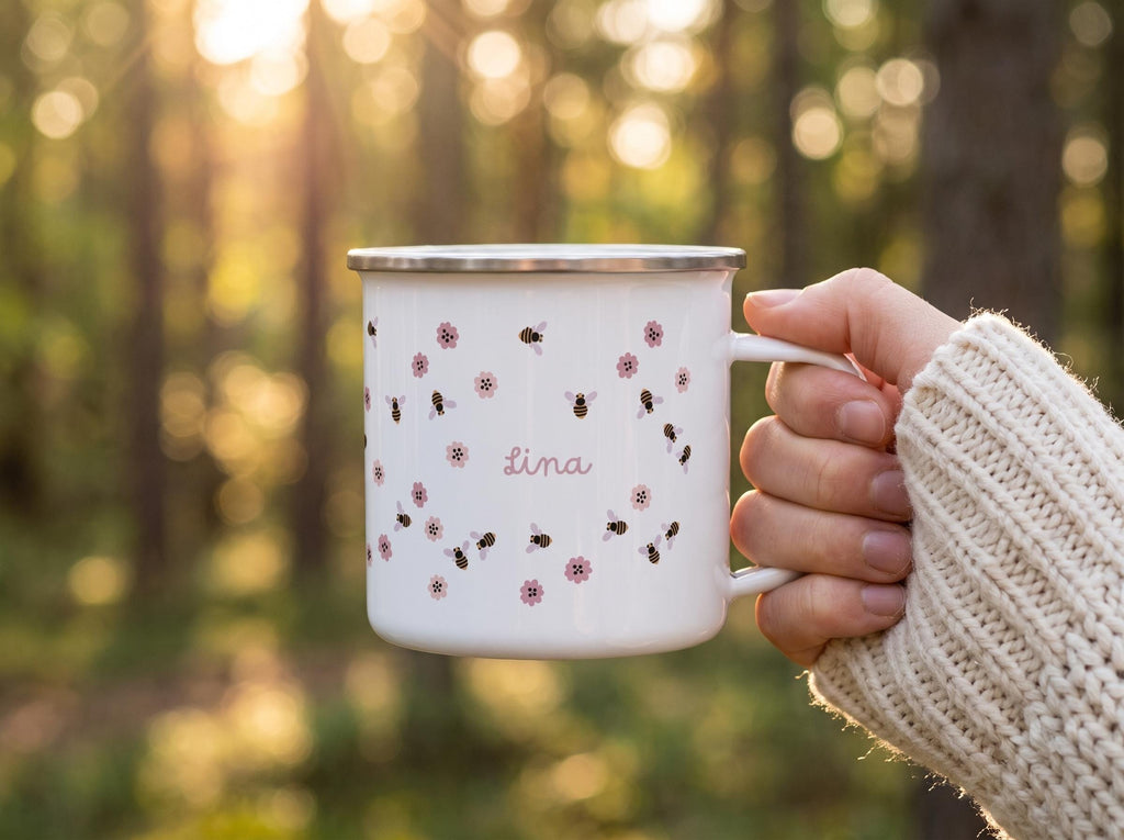 A white enamel mug with a silver rim featuring a repeating pattern of small brown and black bees and pink flowers. The name Lina is printed in a cursive pink font in the centre of the mug. The mug has a single handle on the right side.