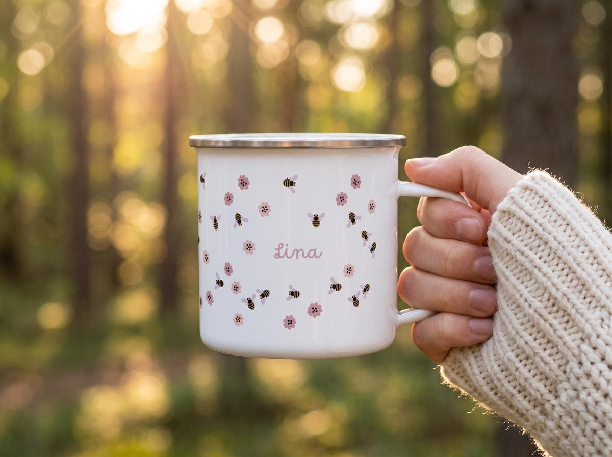 A white enamel mug with a silver rim featuring a repeating pattern of small brown and black bees and pink flowers. The name Lina is printed in a cursive pink font in the centre of the mug. The mug has a single handle on the right side.