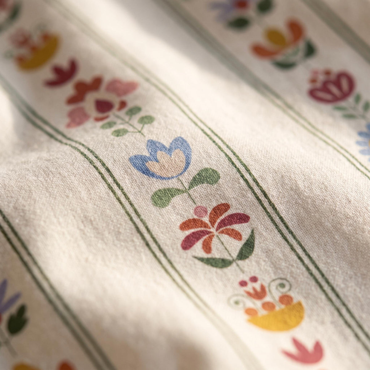 A light beige fabric featuring vertical dark green stripes and a repeating floral pattern. The flowers are printed in various colours including blue, red, orange, and yellow, with green stems and leaves arranged in a linear sequence.