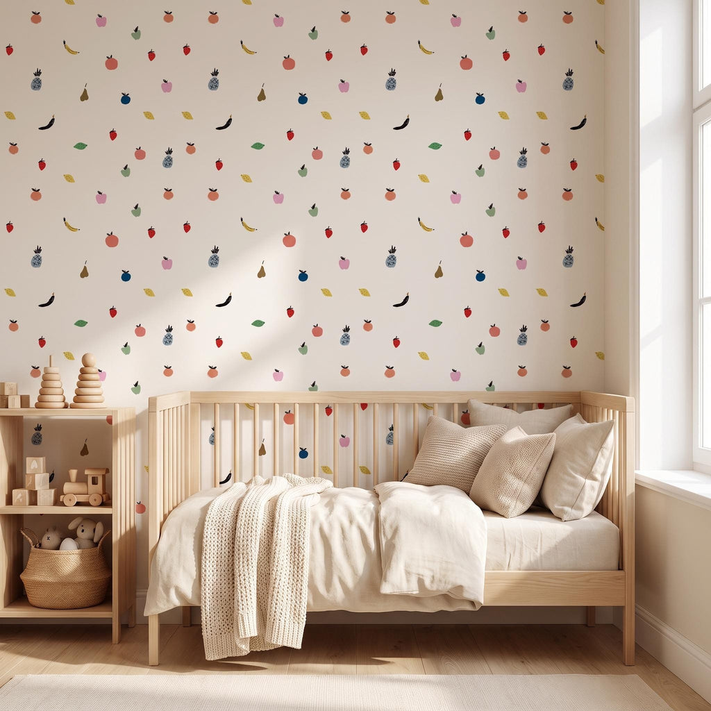 A light-coloured wall featuring a repeating pattern of small, colourful fruit illustrations including pineapples, bananas, strawberries, and apples. A wooden cot with beige bedding and a knit throw blanket sits against the wall.