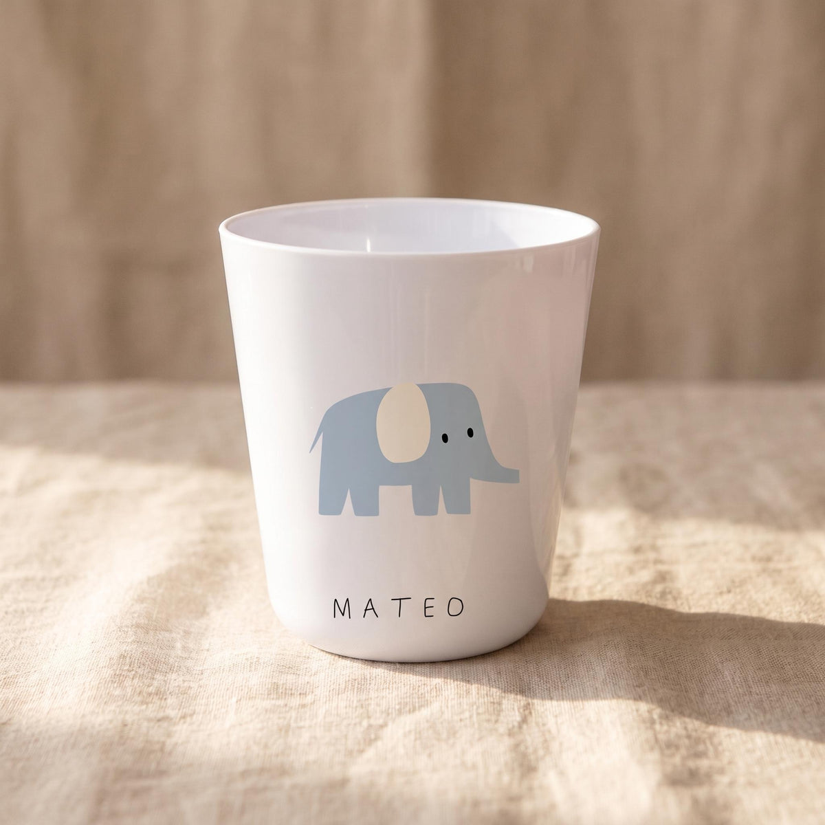 A white tapered cup featuring a light blue elephant graphic with a cream-coloured ear. The name MATEO is printed in black sans-serif capital letters near the base of the cup. The cup has a smooth, glossy finish and a simple, clean design.