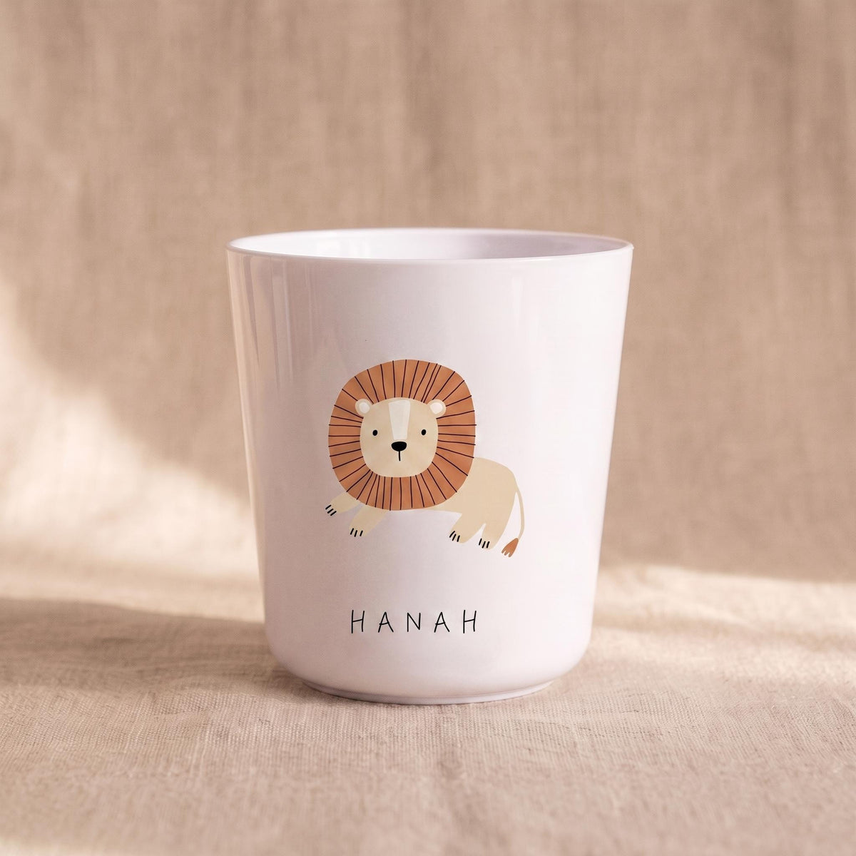 A white cylindrical cup featuring a cartoon lion illustration with a brown mane and a tan body. Below the lion, the name HANAH is printed in simple black capital letters. The cup has a smooth finish and a slightly tapered base.