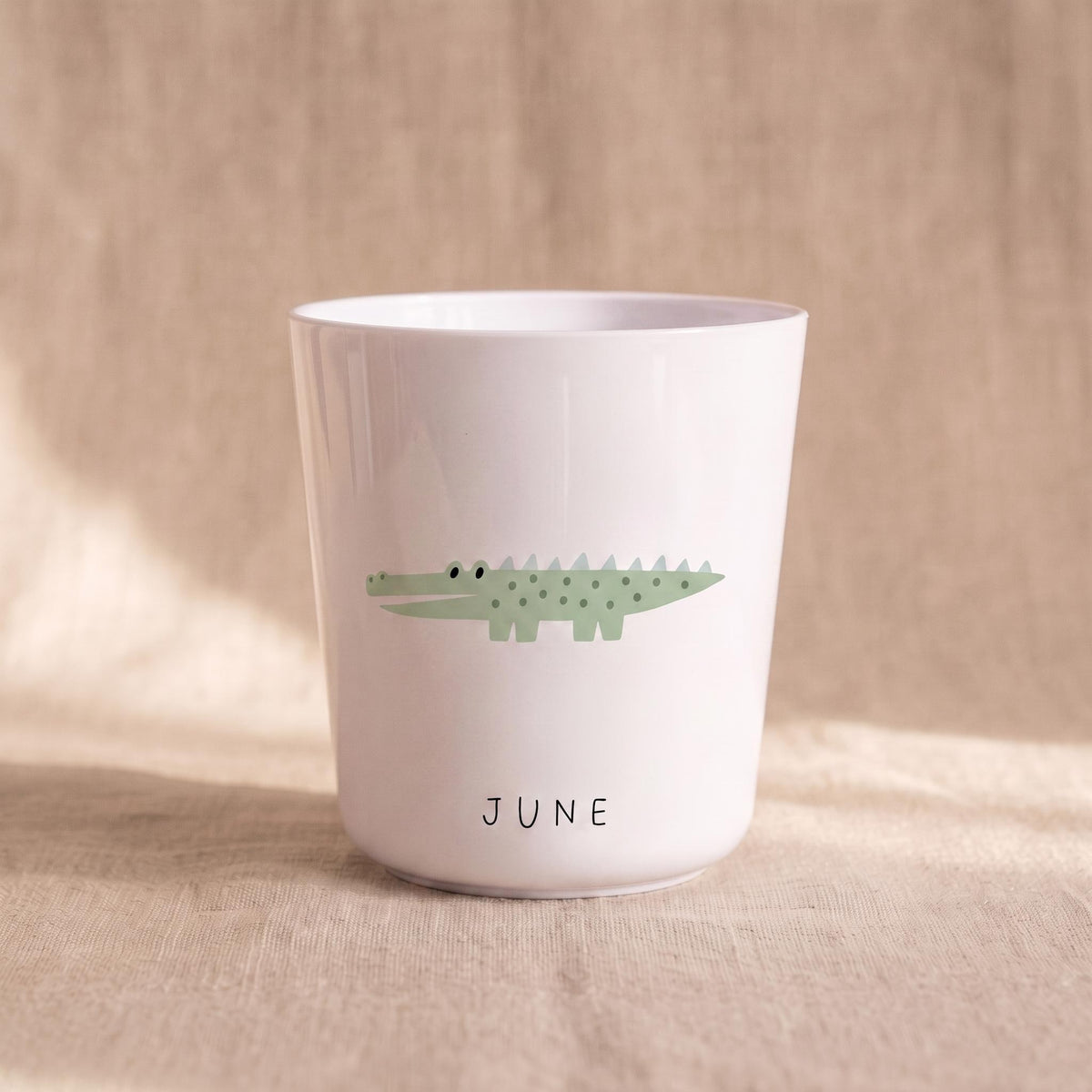 A white cylindrical cup featuring a light green illustration of a crocodile with dark green spots and a row of spikes on its back. The word JUNE is printed in simple black capital letters below the crocodile graphic on the cup.