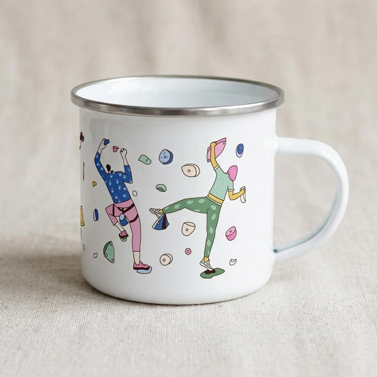 A white enamel mug featuring a climber design rests on a neutral surface.