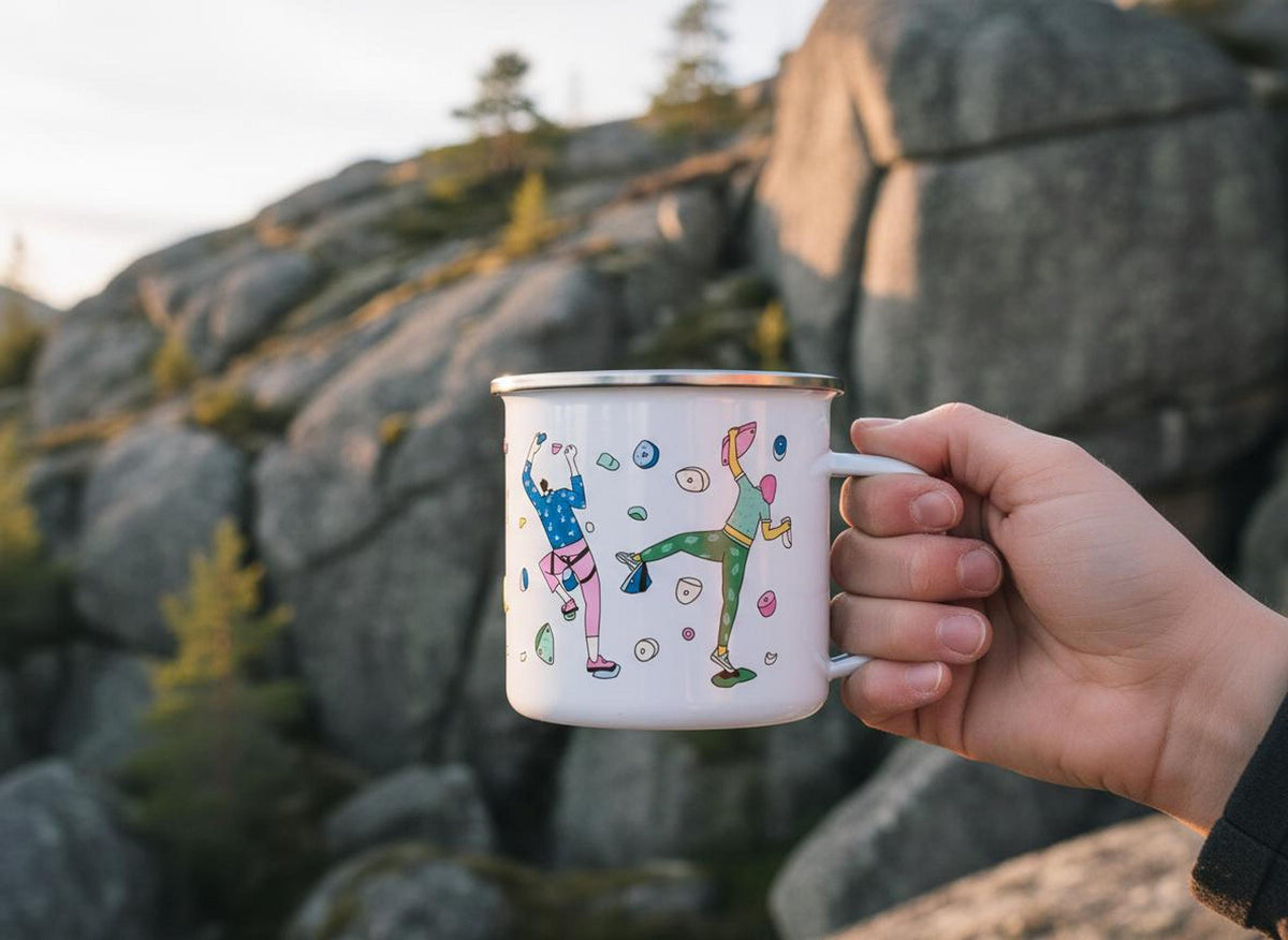 A hand holds a white enamel climbing mug in front of rocky boulders.