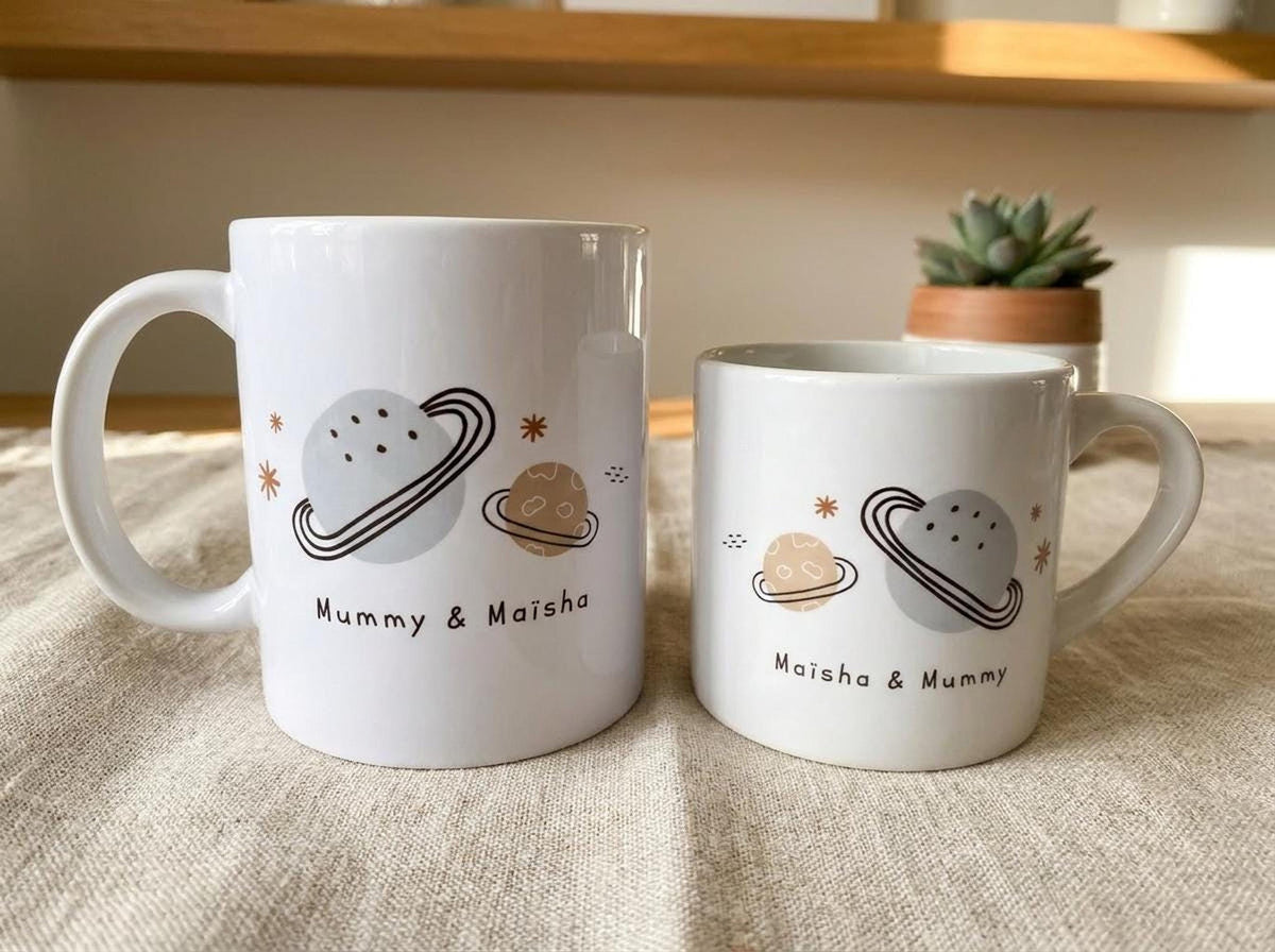 Two white ceramic mugs of different sizes sit on a textured cloth surface, featuring printed space graphics of planets and the names Mummy and Maisha.