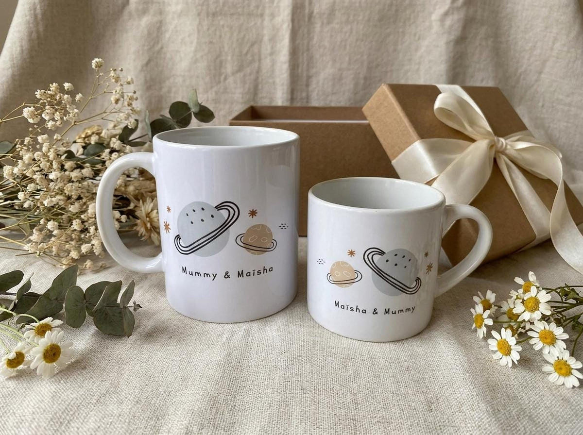 Two matching white mugs sit alongside a gift box tied with a satin ribbon, dried flowers, and small white daisies on a neutral fabric.