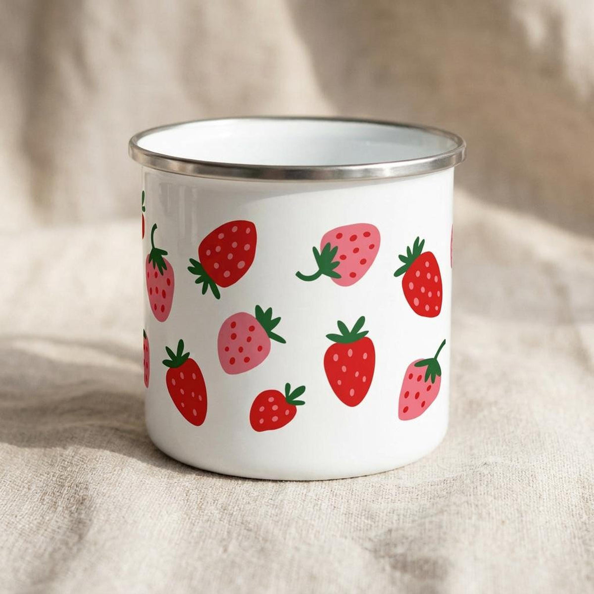 A white enamel mug with a silver rim displays a scattered strawberry pattern against a neutral textured background.