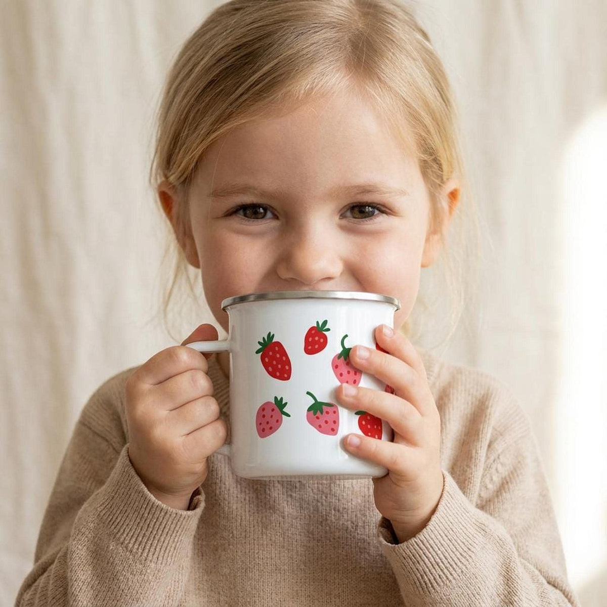 A young girl with blonde hair takes a sip from a white enamel mug printed with small red and pink strawberries.
