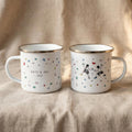 Two white enamel camping mugs featuring rock climbing graphics sit side by side on a neutral fabric background.