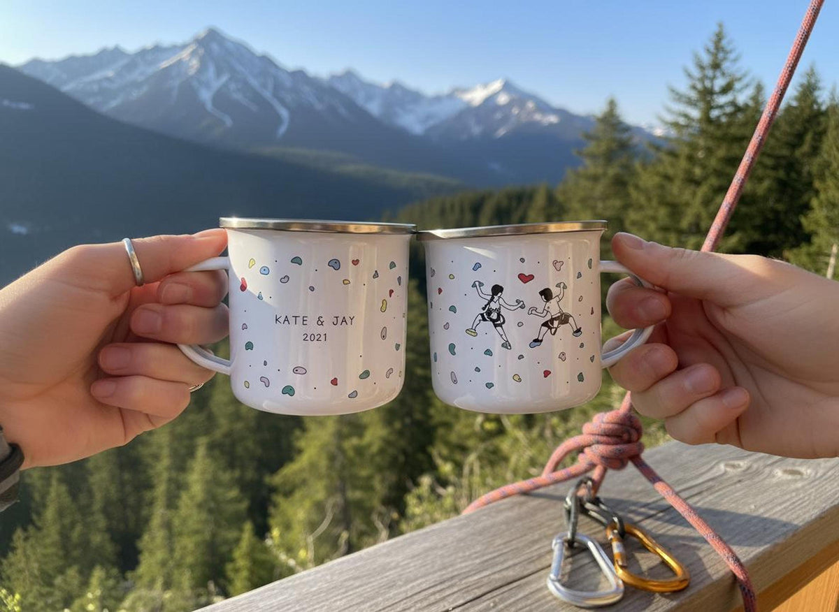 Two hands hold two white enamel mugs together on a wooden railing overlooking mountains, with climbing gear nearby.