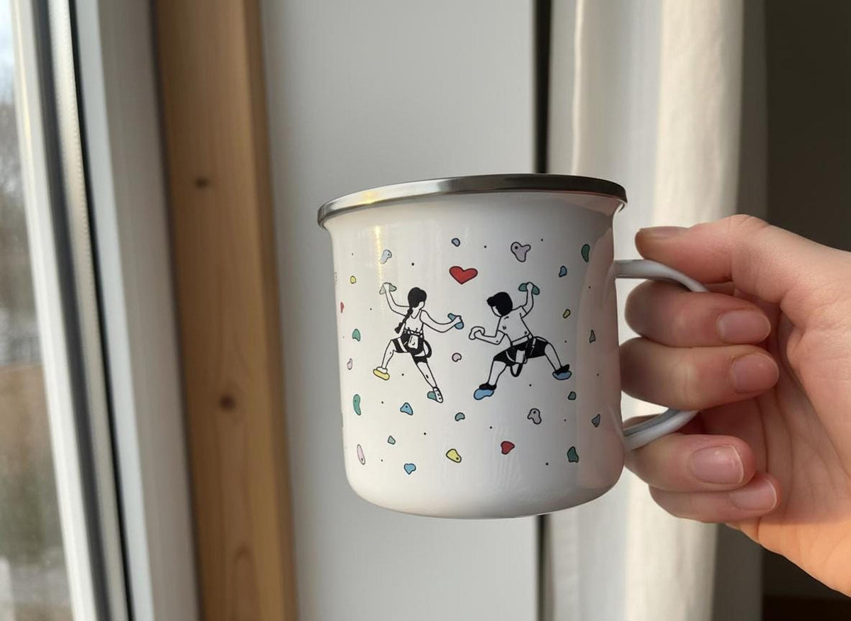 A hand holds a white enamel mug with a rock climbing illustration in front of a bright window.