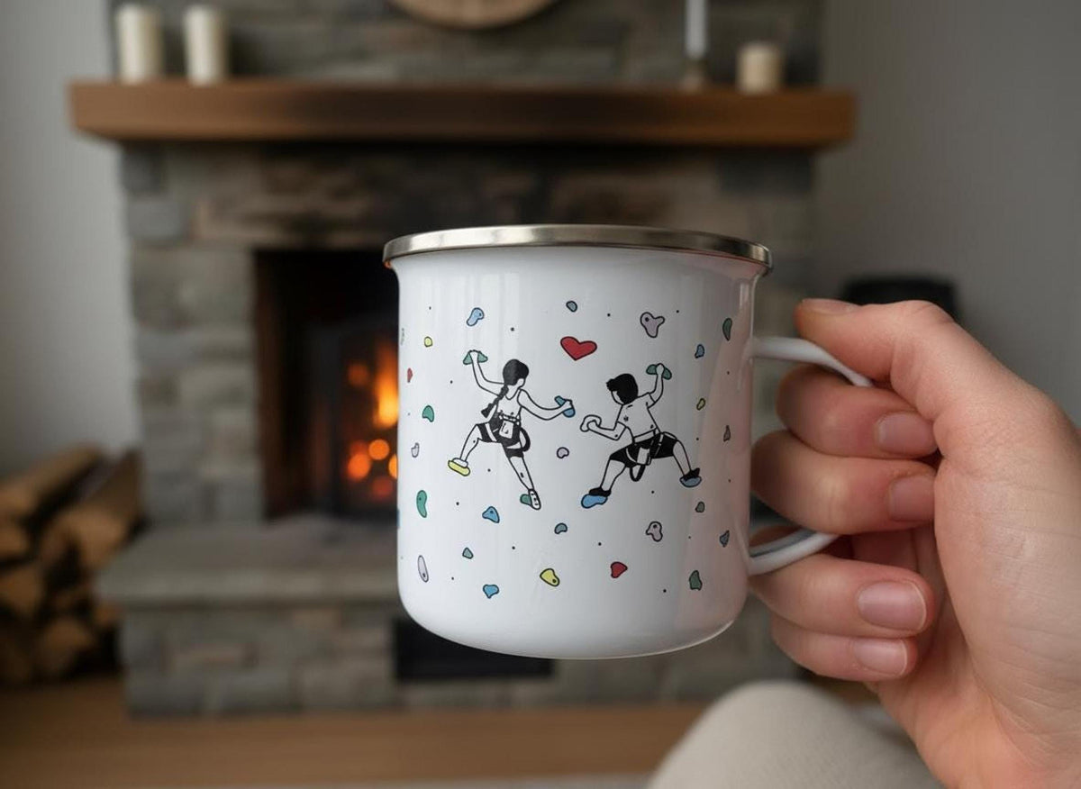 A hand holds a white enamel mug printed with a rock climbing couple illustration in front of a stone fireplace.
