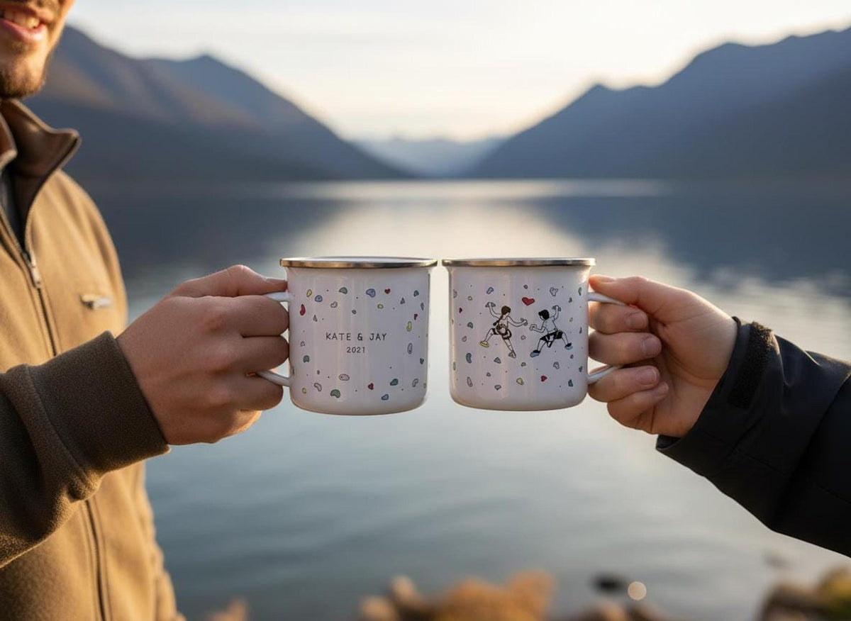 Two people hold up two white enamel camping mugs against a scenic lake and mountain background during sunset.