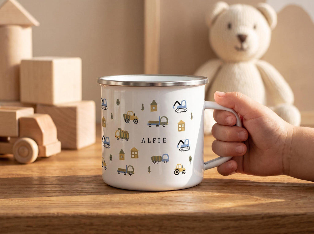 A child's hand holds a white enamel mug featuring a pattern of construction vehicles, trees, and houses, with the name ALFIE printed in the center.