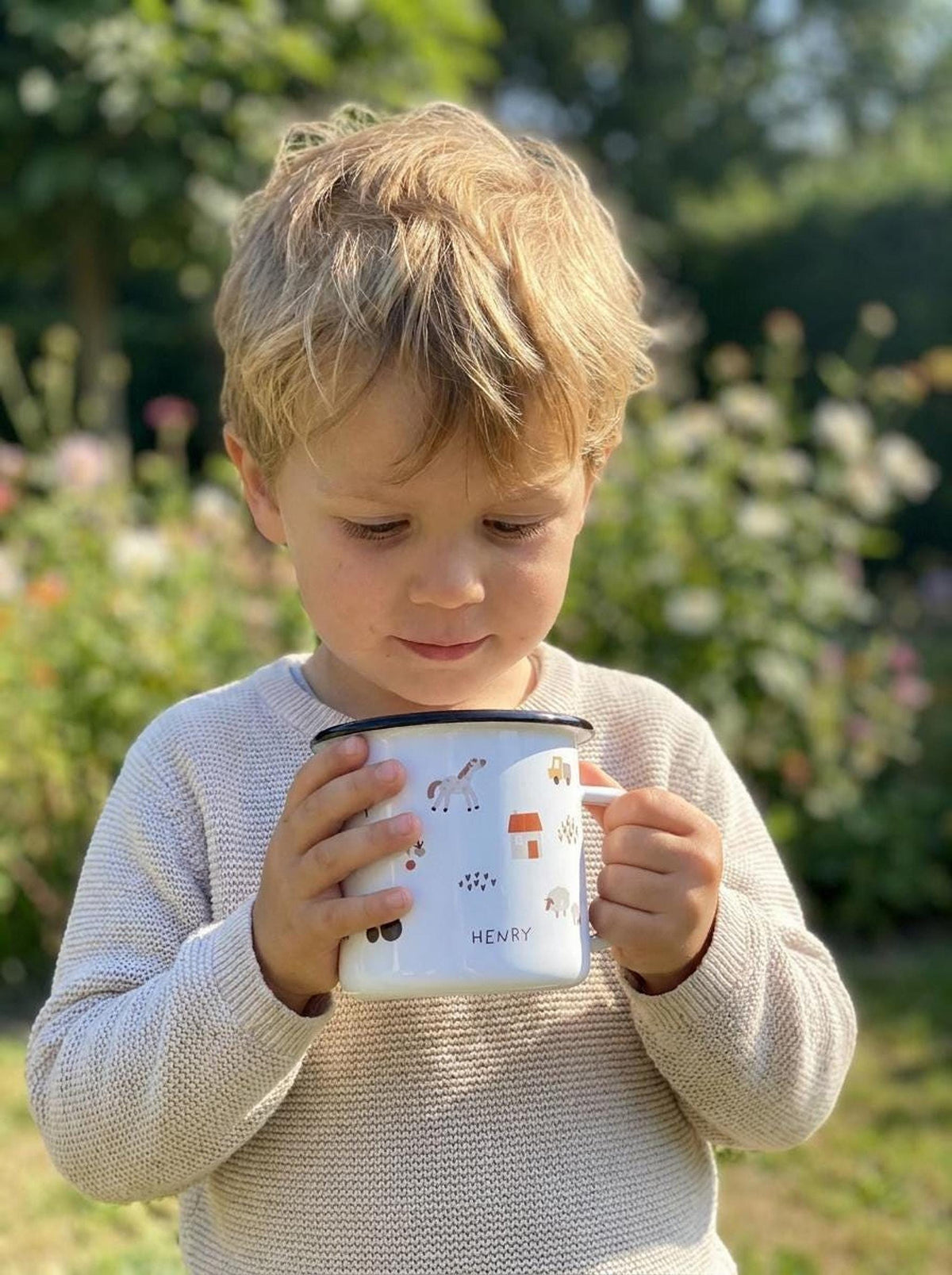 A young child holding a personalized enamel mug with farm illustrations outdoors in a garden.