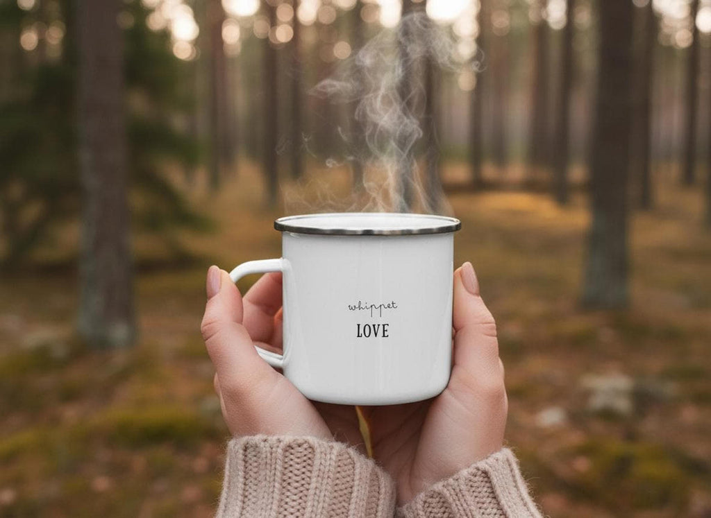 Hands in a knitted sweater hold a steaming white enamel mug printed with the text whippet LOVE in front of a blurred forest background.