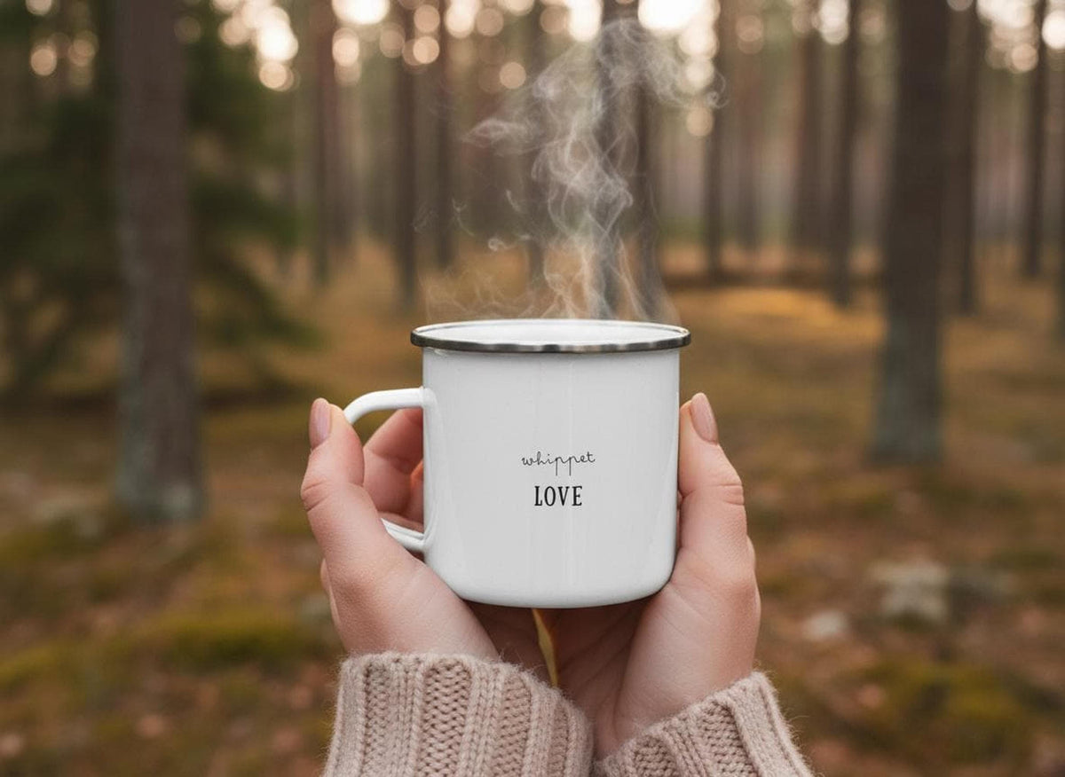 Hands in a knitted sweater hold a steaming white enamel mug printed with the text whippet LOVE in front of a blurred forest background.