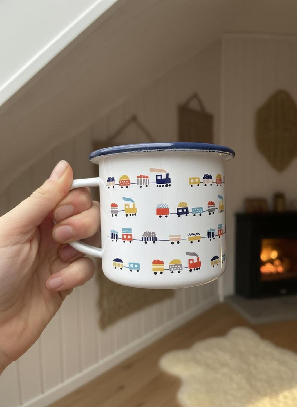 A hand holds a white enamel mug with a blue rim and a colorful train print against an interior backdrop with a fireplace.
