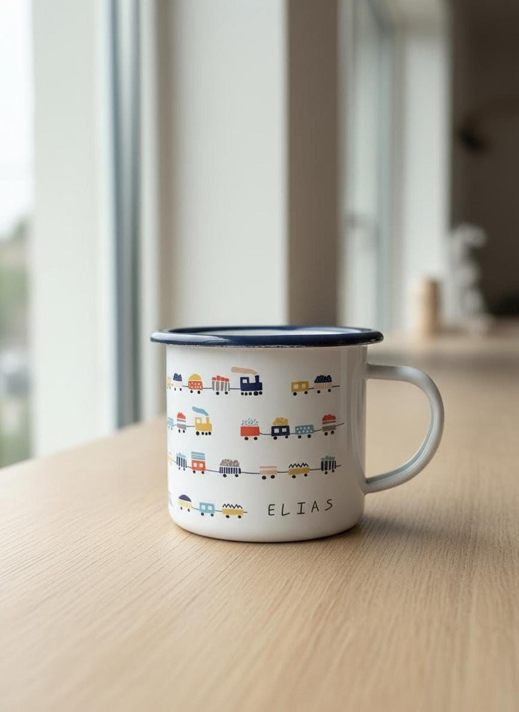 A personalised white enamel mug with a blue rim and train pattern showing the name Elias, placed on a light wooden surface by a window.