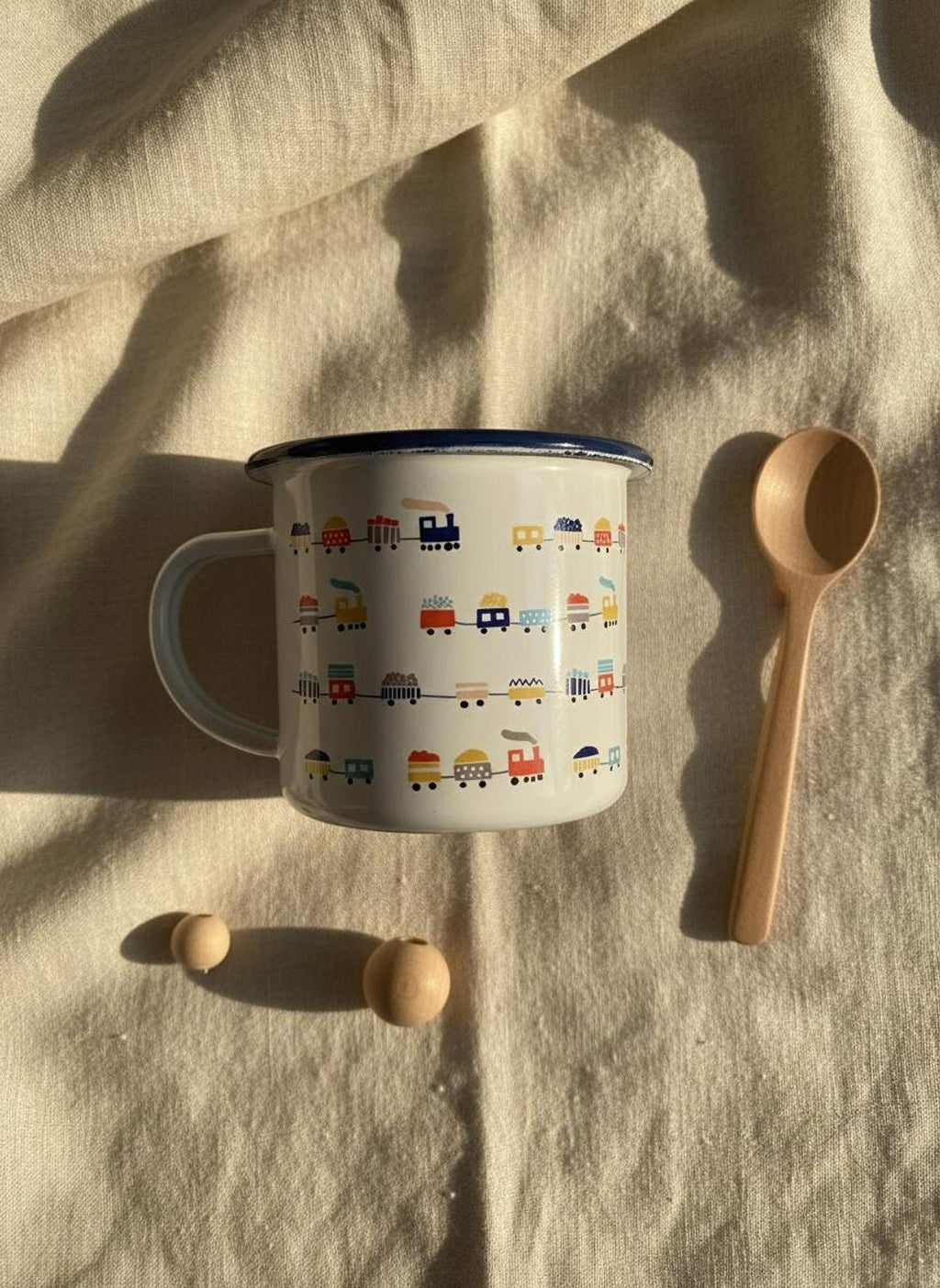 A white enamel mug with a train print and a blue rim rests on a beige fabric cloth alongside a wooden spoon and two wooden beads.
