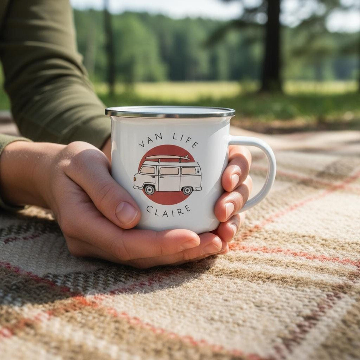 A person holds a white enamel mug featuring a red campervan and the name Claire on a blanket outdoors.