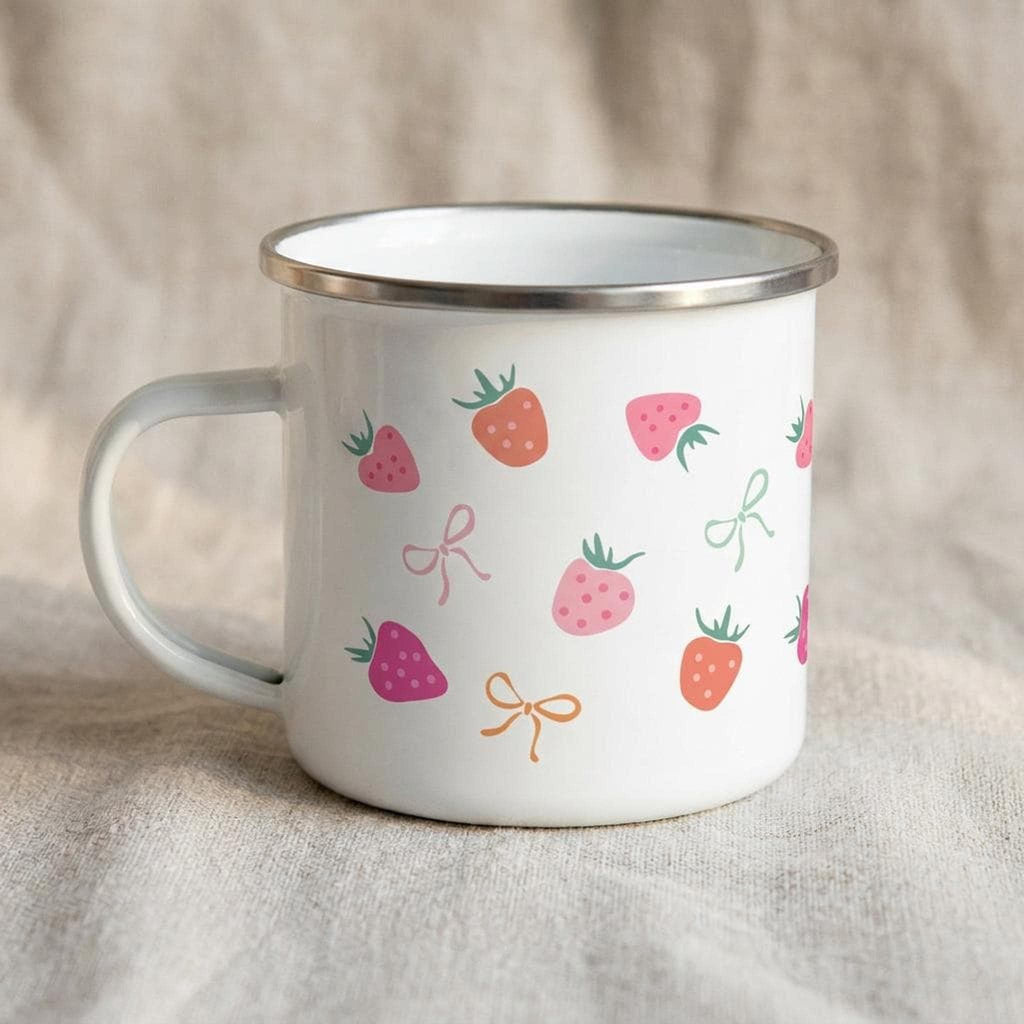 A white enamel mug featuring a silver rim and a pattern of pink and red strawberries and pastel bows, positioned on a textured beige surface.