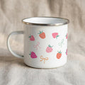 A white enamel mug featuring a silver rim and a pattern of pink and red strawberries and pastel bows, positioned on a textured beige surface.