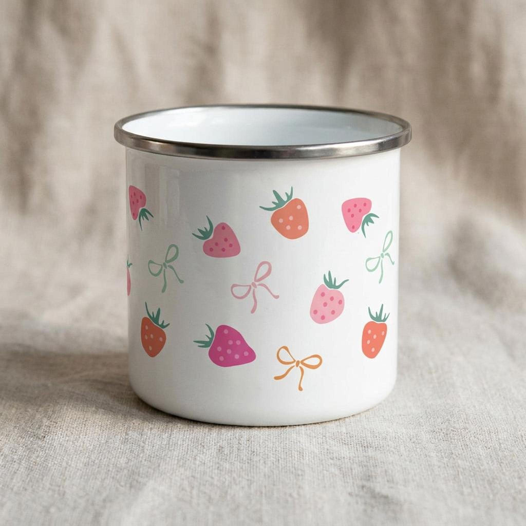 A white enamel mug with a silver rim, decorated with a repeating pattern of strawberries and small bows in pink, red, and green on a neutral fabric.