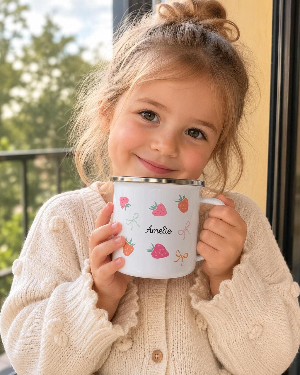 A smiling young girl in a knitted cream sweater holds a personalised white enamel mug with a strawberry pattern up to her cheek.