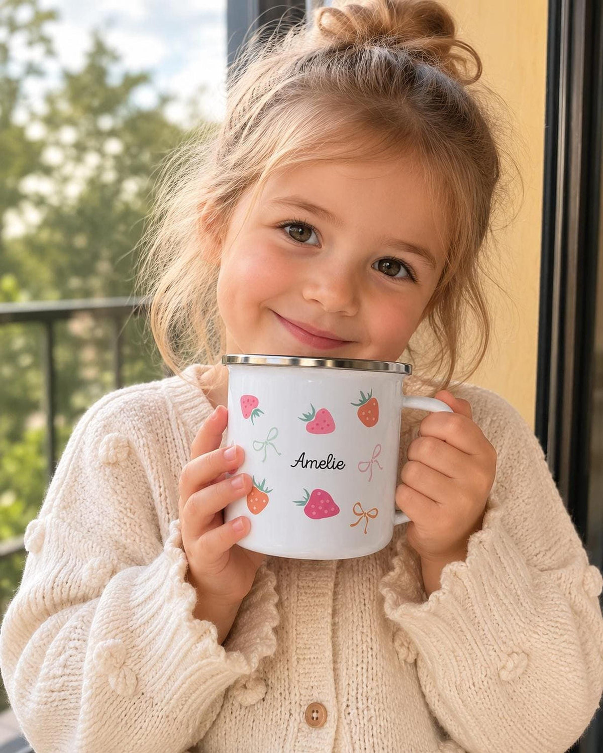 A smiling young girl in a knitted cream sweater holds a personalised white enamel mug with a strawberry pattern up to her cheek.