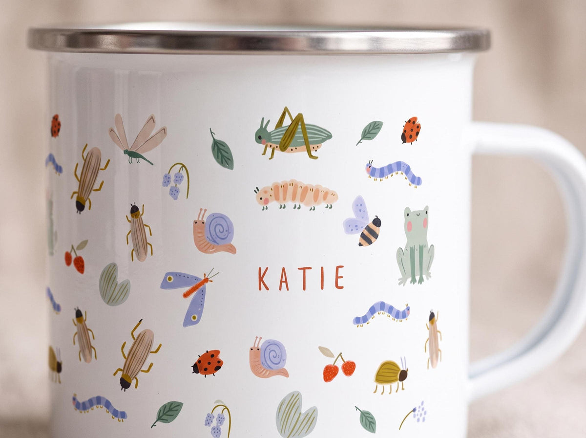 Close-up of a white enamel mug featuring colorful illustrations of insects and the name KATIE in orange letters.