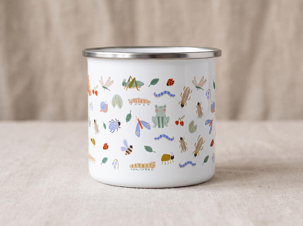 White enamel mug with a silver rim, covered in small colorful bug illustrations, placed on a light beige cloth.