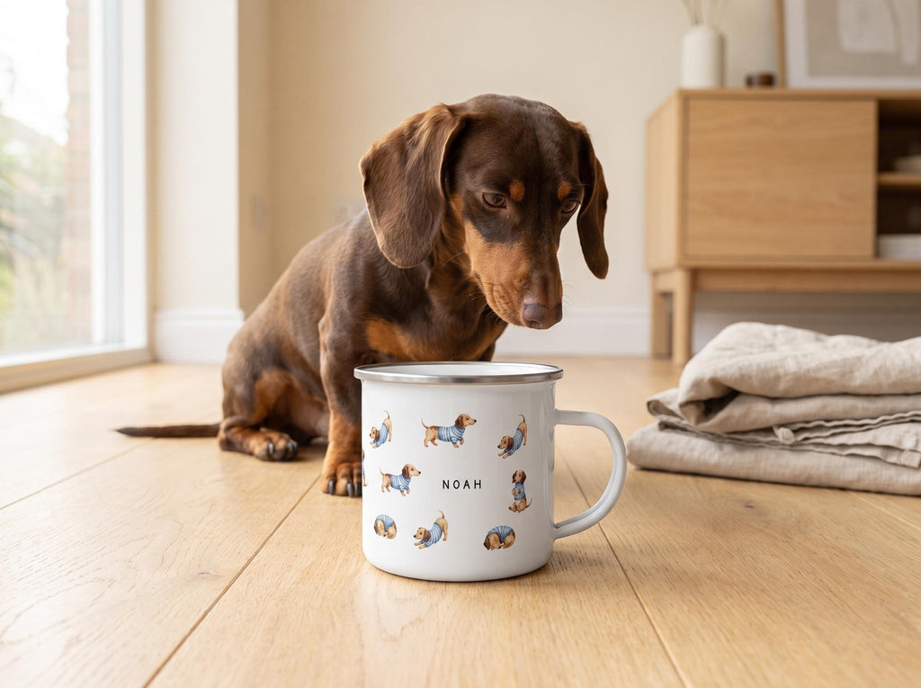 A real brown dachshund sits on a wooden floor next to a white enamel mug printed with illustrations of dachshunds and the name Noah.
