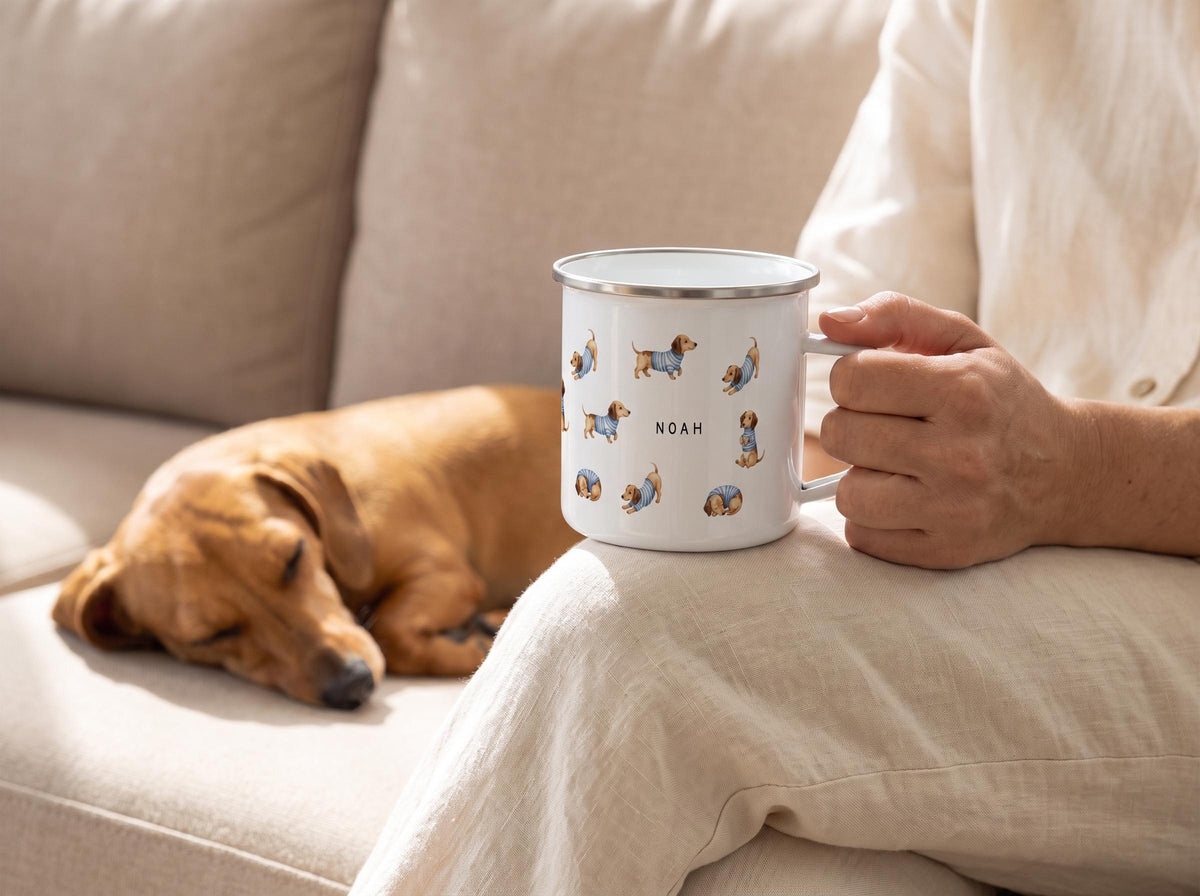 A person in cream loungewear holds a personalized dachshund enamel mug, while a real dachshund sleeps on the sofa next to them.