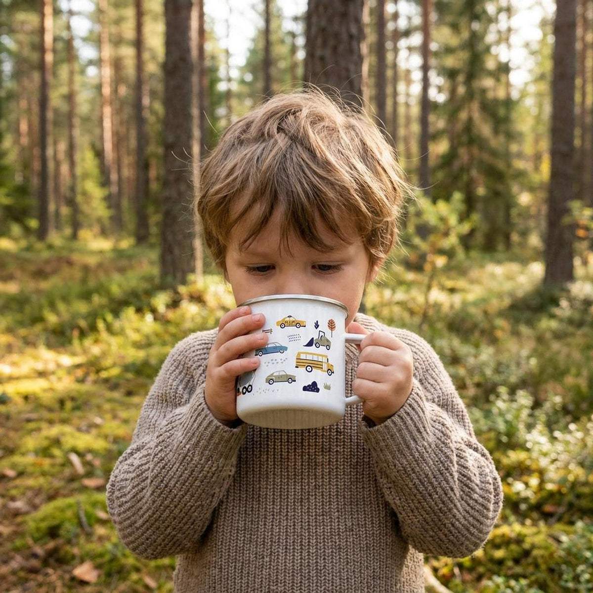 A young child wearing a beige knit sweater drinks from a white enamel mug with transport vehicle illustrations in a forest.