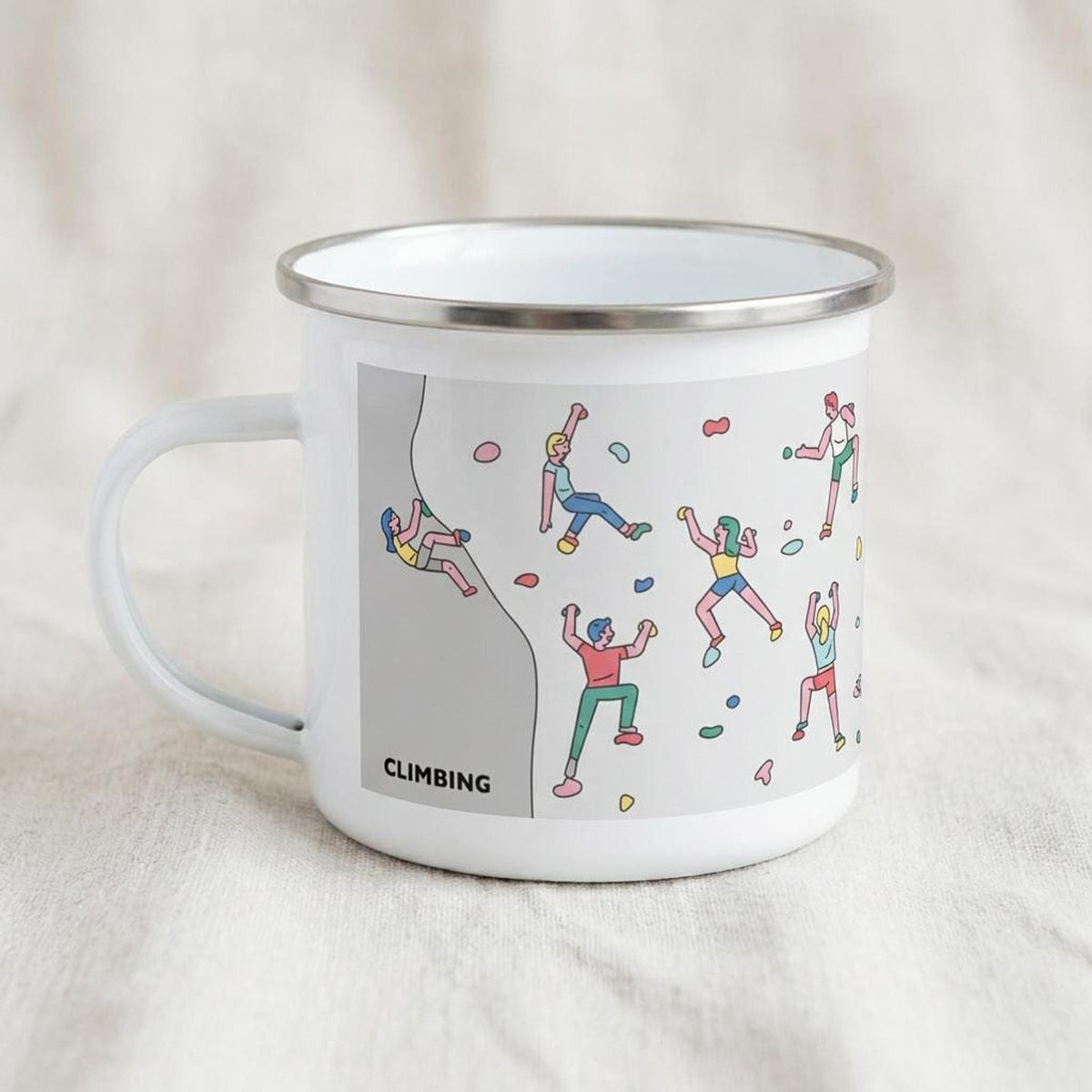 A white enamel mug with a silver rim sits on a textured light surface, featuring an illustration of rock climbers and the word CLIMBING.