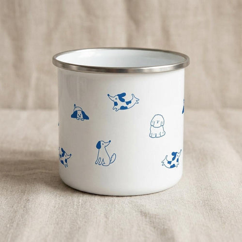 A white enamel mug with a silver rim displays various blue dog illustrations across its side, shown against a neutral fabric background.