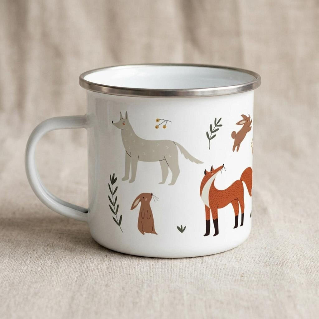 A white enamel mug with a silver rim sits on a beige fabric surface, featuring illustrations of a gray wolf, a red fox, rabbits, and foliage.