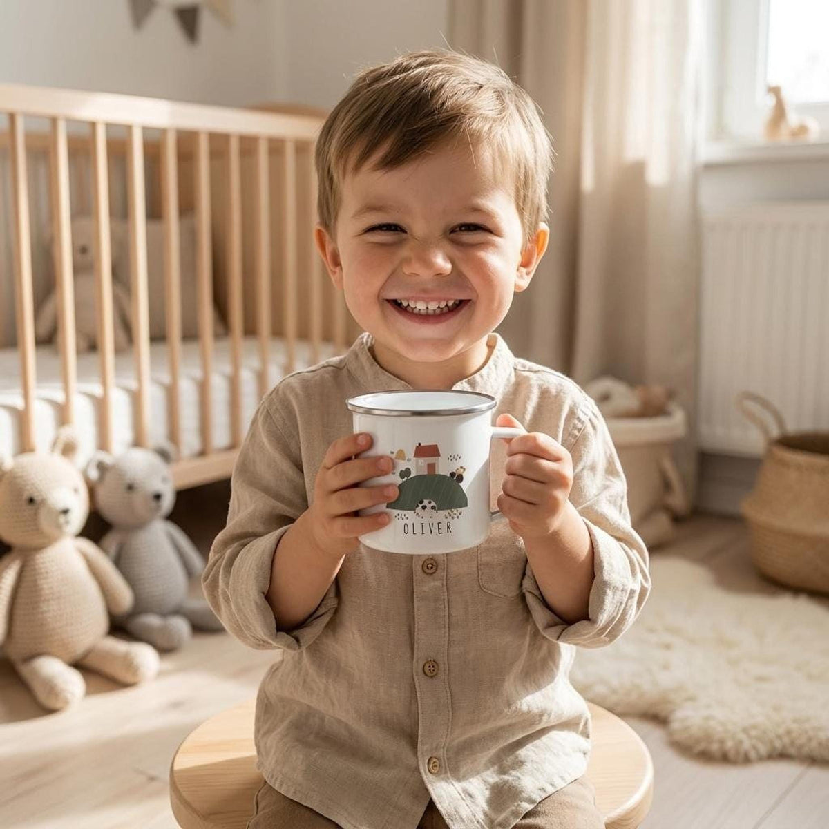 A smiling young child in a tan shirt holds a white enamel mug with a farm design in a nursery with stuffed animals and a crib.
