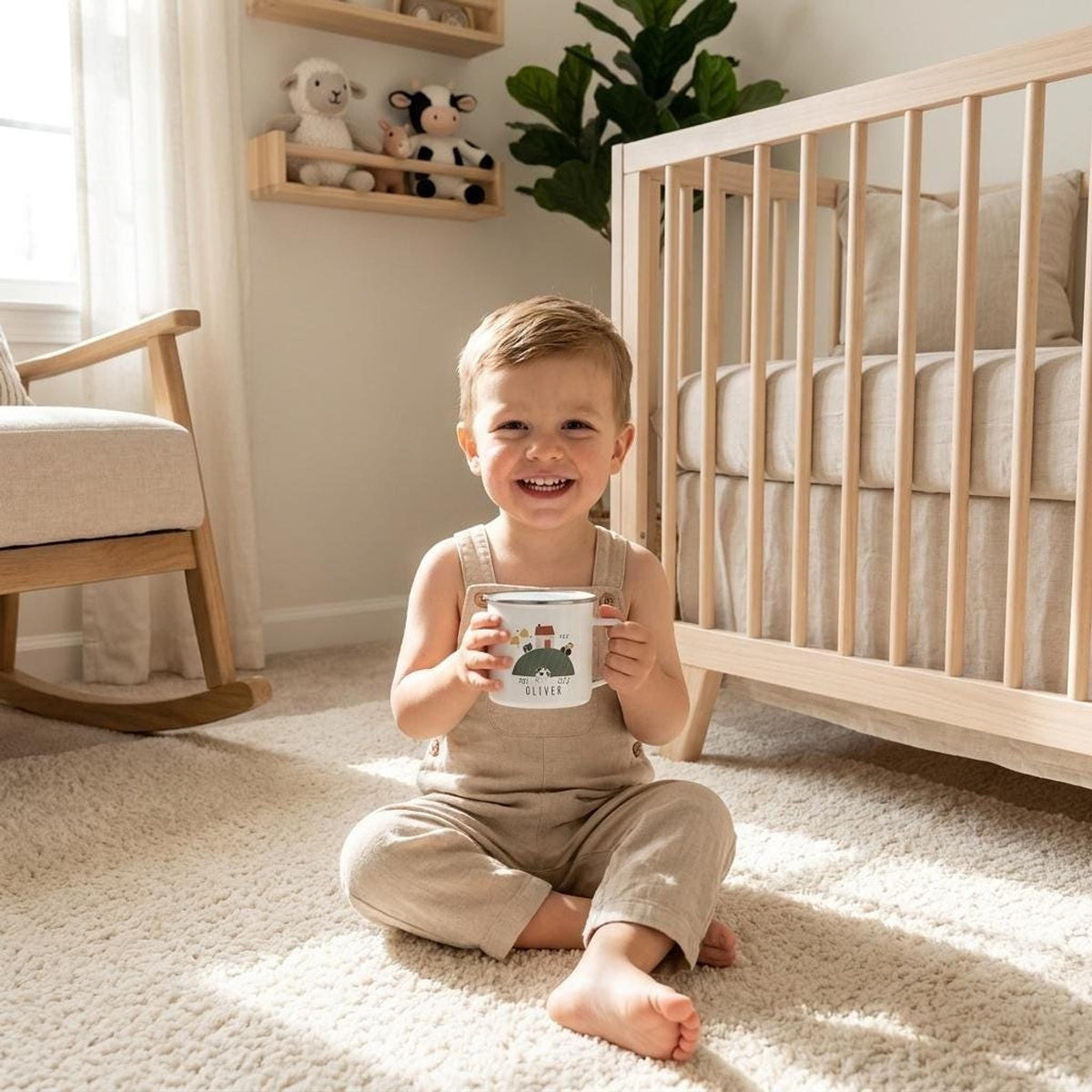 A toddler in tan overalls sits on a rug in a brightly lit room holding a white enamel mug with a farm graphic in front of a crib.