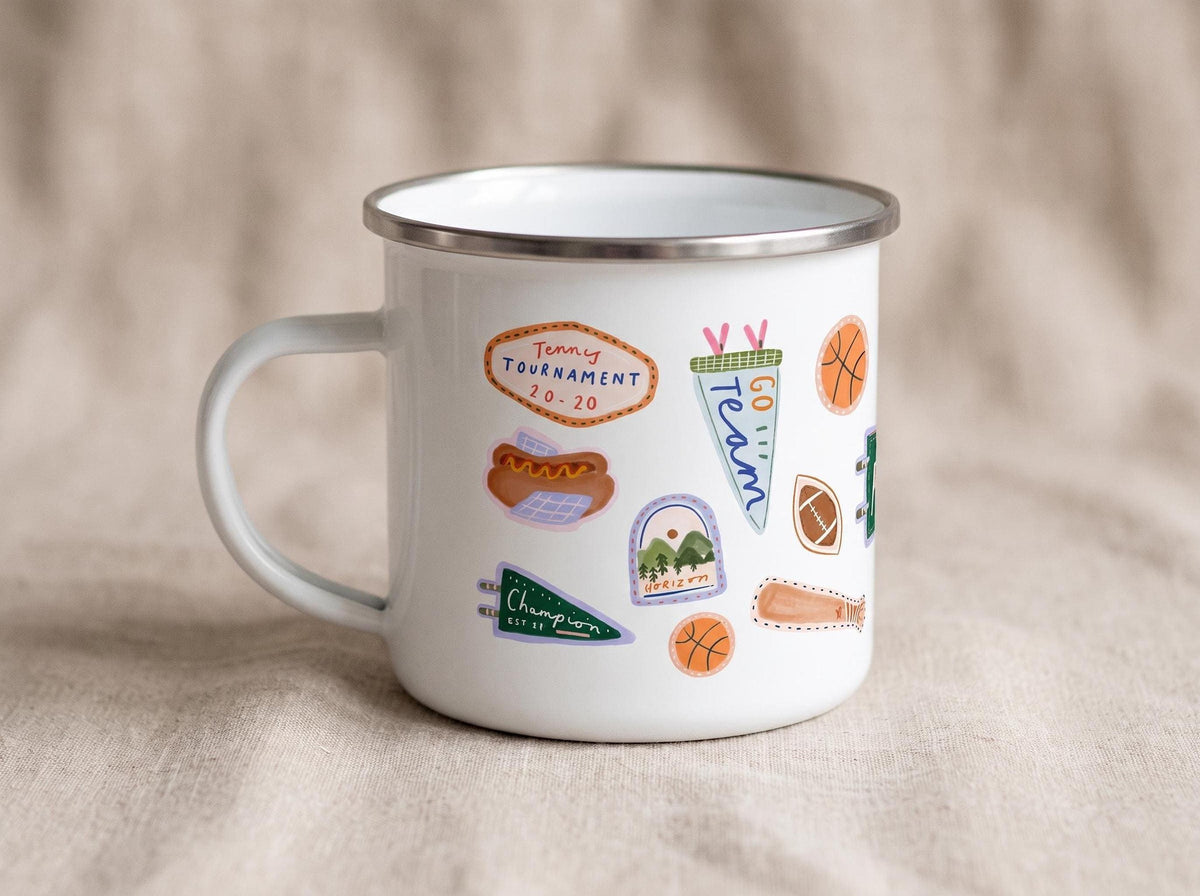 A white enamel mug with a silver rim, featuring various colourful sports-themed stickers including a tennis tournament graphic and a green pennant.